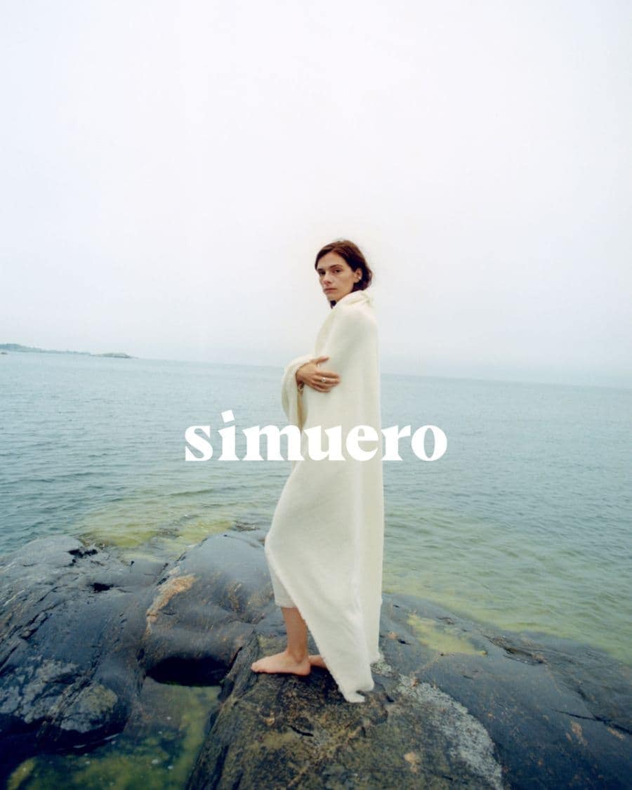 Model wrapped in a white blanket standing on dark coastal rocks by the sea under an overcast sky for Simuero.