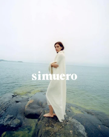 Model wrapped in a white blanket standing on dark coastal rocks by the sea under an overcast sky for Simuero.