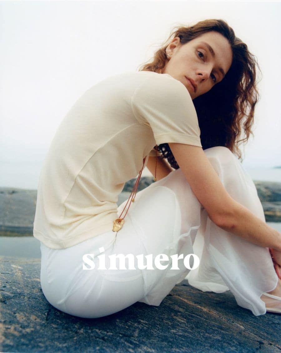 Model wearing a light cream short-sleeve top and white trousers sitting on rocky coastal terrain