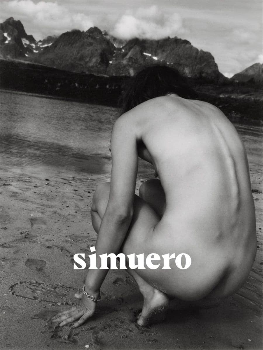 Monochrome photograph of a person crouching on a wet sandy beach with a mountain range rising across the water in the background.