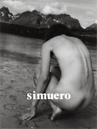 Monochrome photograph of a person crouching on a wet sandy beach with a mountain range rising across the water in the background.