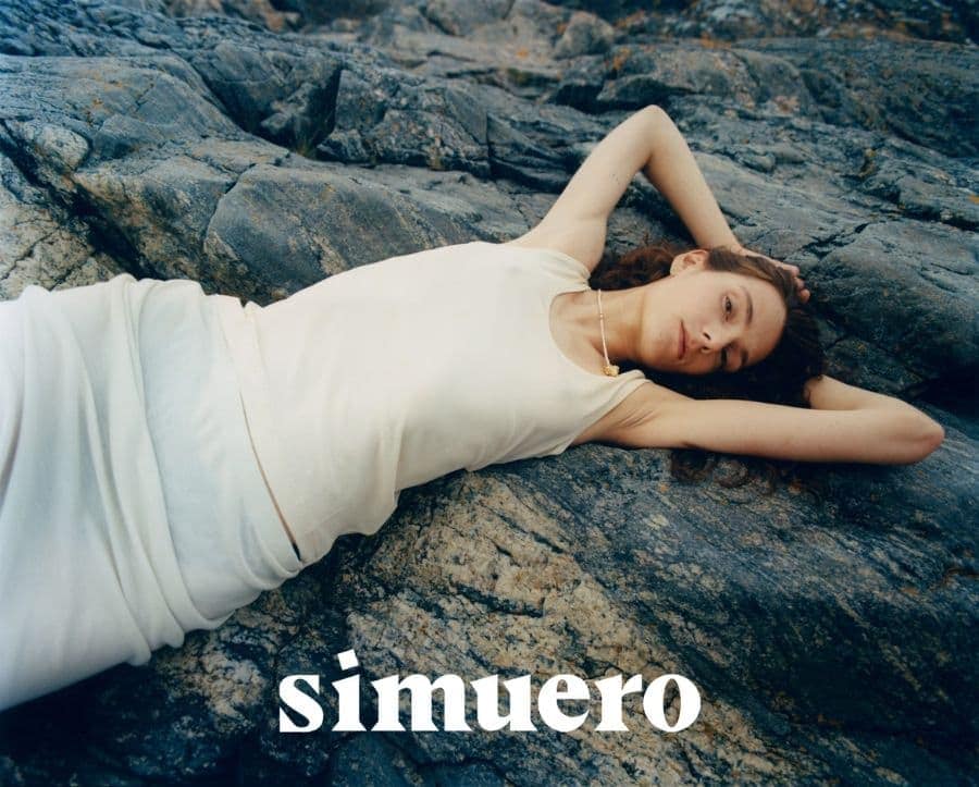 Model lying on dark rocky coastal ground wearing a cream white tank top for Simuero Marea Collection 2024 campaign in Lofoten Norway