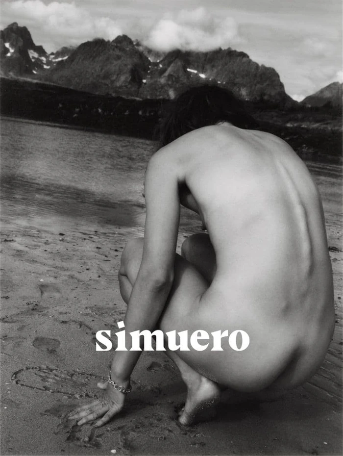 Monochrome photograph of a person crouching on a wet sandy beach with a mountain range rising across the water in the background.
