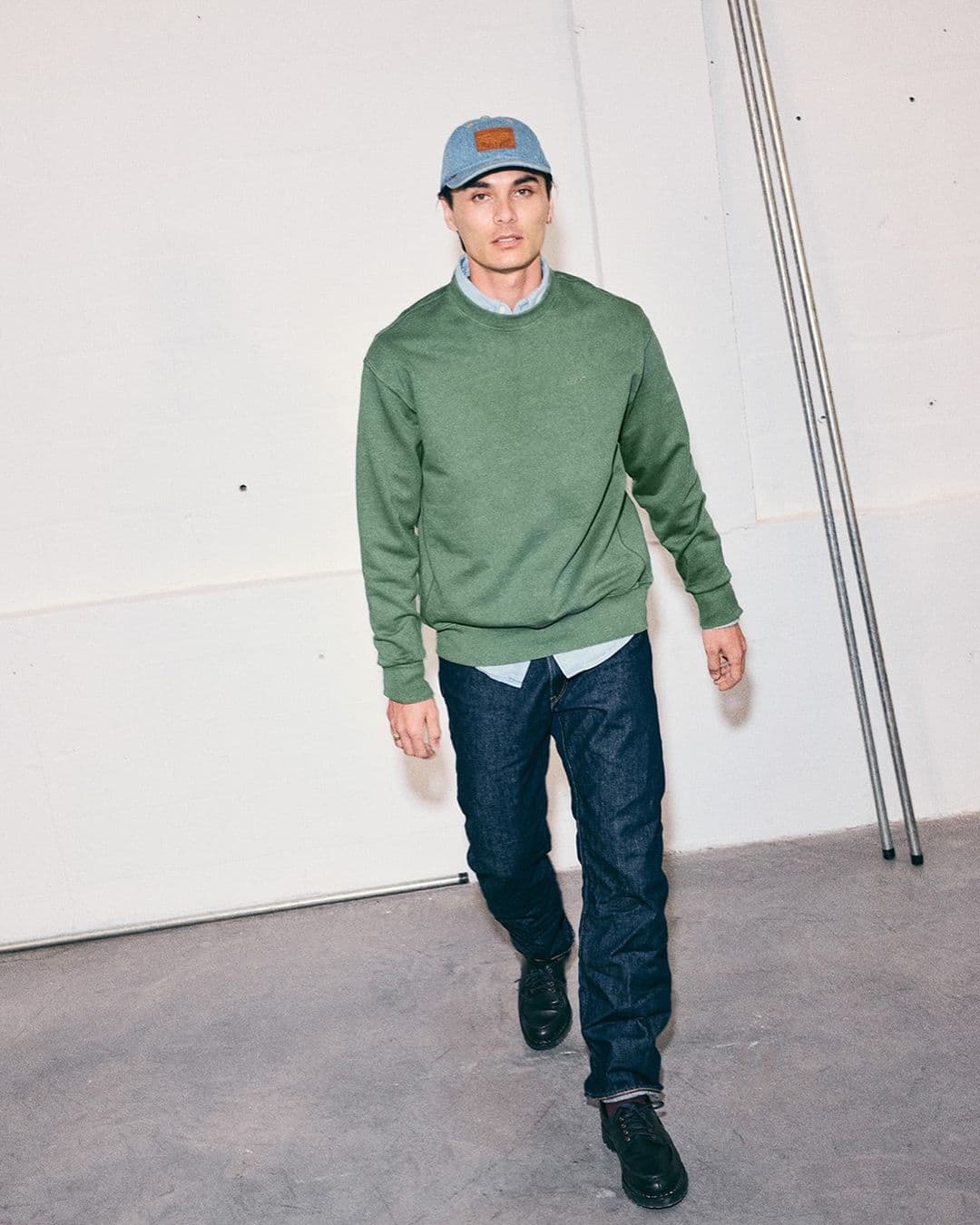 Full-length portrait of a man wearing a green sweater, blue jeans, and cap walking forward in front of a white wall.