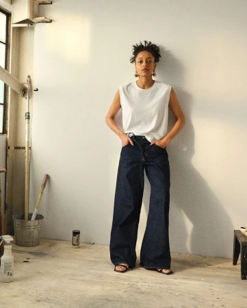Model standing against a plain wall wearing a white sleeveless top and dark denim jeans in a bright studio.