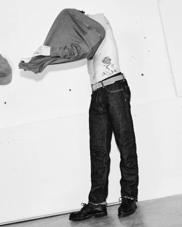 Man standing against a white wall pulling off his grey sweatshirt, wearing dark denim jeans and black shoes.