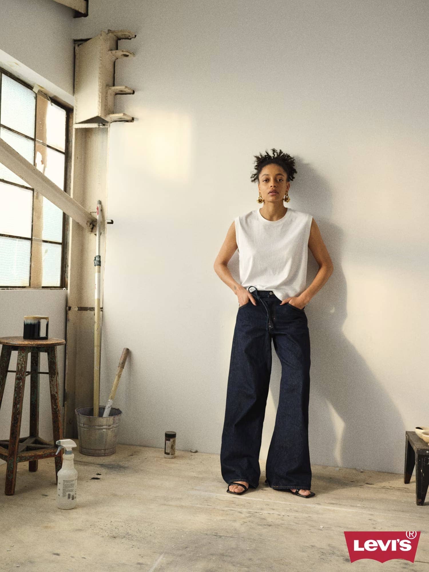 Model wearing a white tank top and dark denim jeans leaning against a plain wall in a light-filled studio space.