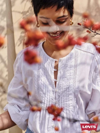 Smiling person wearing a white button-front blouse with lace details, framed by blurred red berries in the foreground.