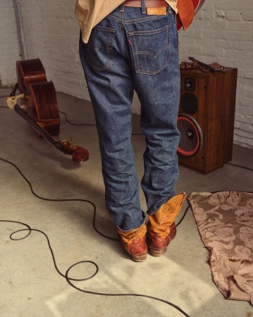 Rear view of a person wearing blue denim jeans and tan cowboy boots standing on a concrete floor in a room with a wooden speaker and double bass.