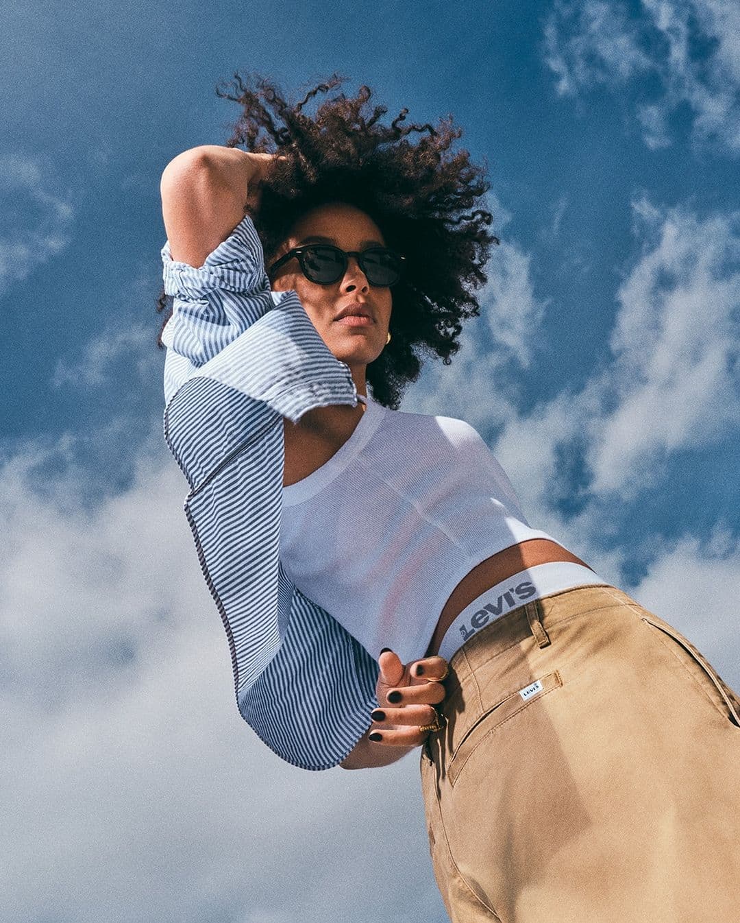 Model with dark curly hair wearing sunglasses, white tank top, and khaki pants against a blue sky