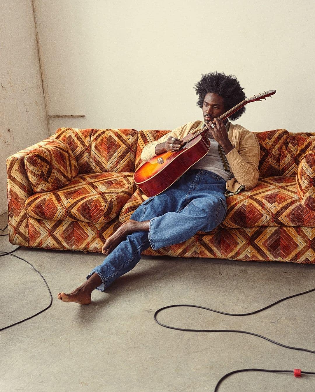 Man with afro sitting barefoot on a patterned vintage sofa playing an acoustic guitar indoors.