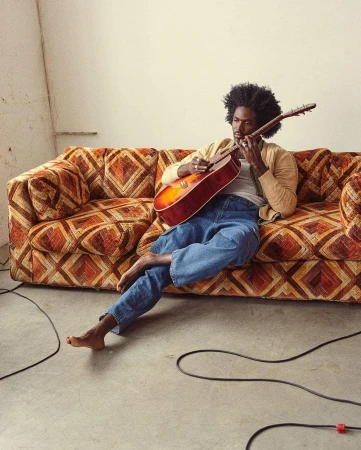 Man with afro sitting barefoot on a patterned vintage sofa playing an acoustic guitar indoors.