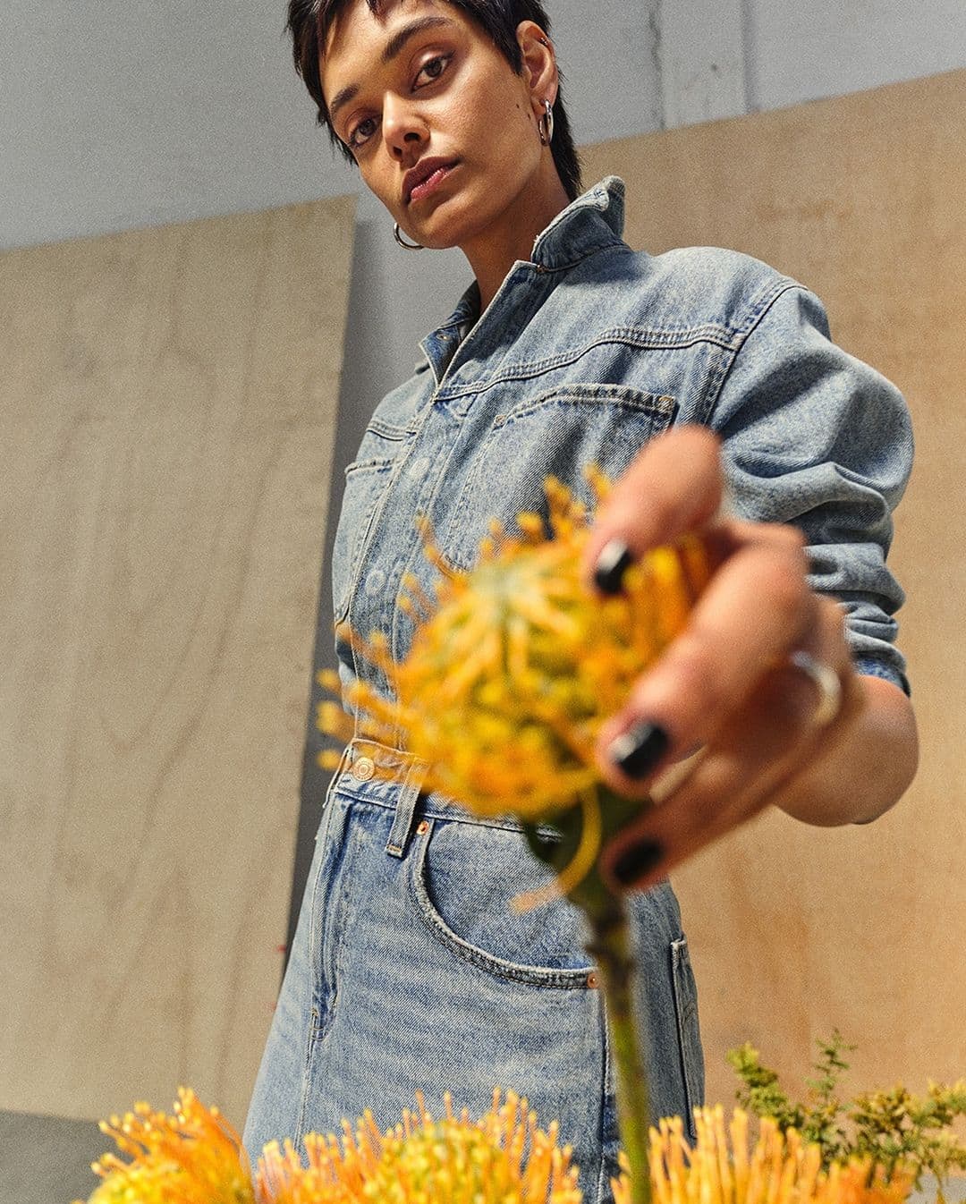 Model in denim outfit holding a yellow flower towards the camera against wooden panels in Levi's Back At It ad campaign.
