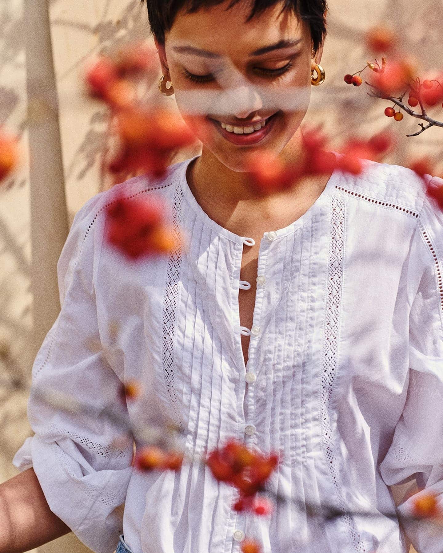 Smiling person wearing a white button-up blouse with red berries framing the foreground in natural light.