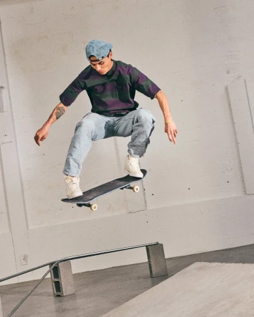 Skater mid-air performing a trick over a rail inside an industrial indoor space wearing a striped t-shirt and light jeans