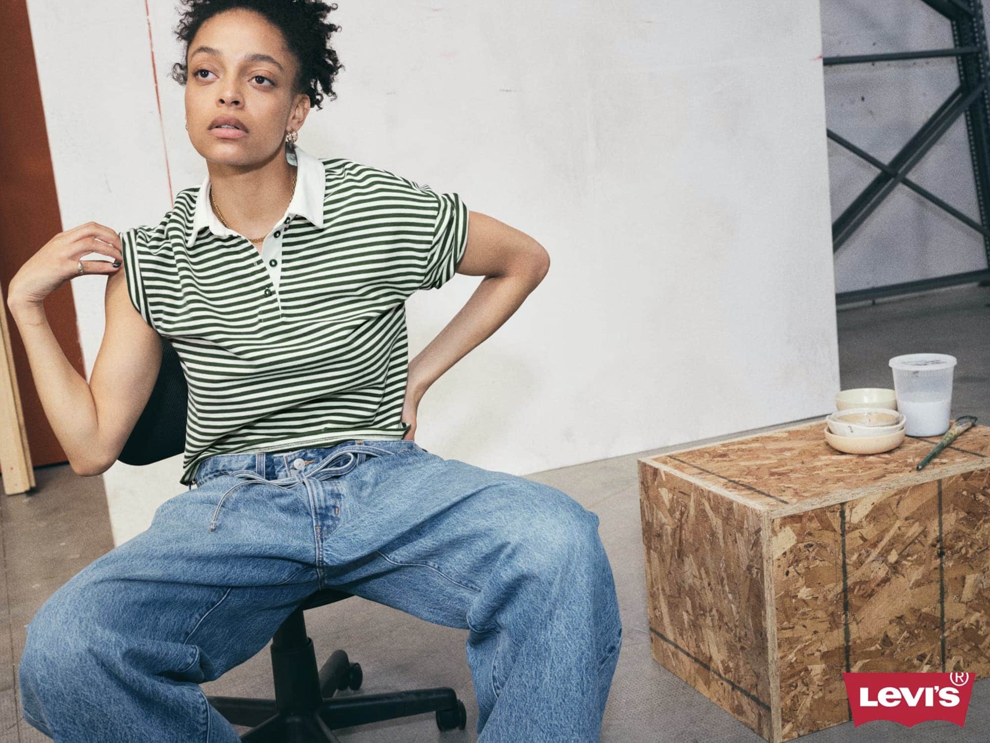 Woman seated on an office chair wearing a green and white striped polo shirt and loose denim jeans.