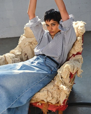 Model with short dark hair in a striped button-down shirt and long denim skirt seated in an armchair