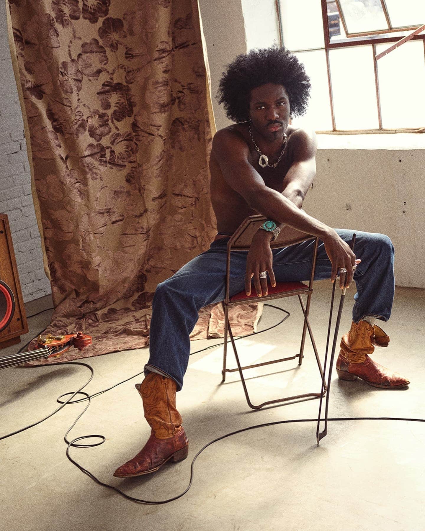 Shirtless man with afro sitting backward on folding chair wearing denim jeans and cowboy boots