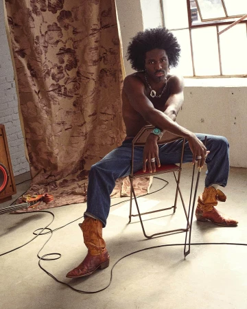 Shirtless man with afro sitting backward on folding chair wearing denim jeans and cowboy boots