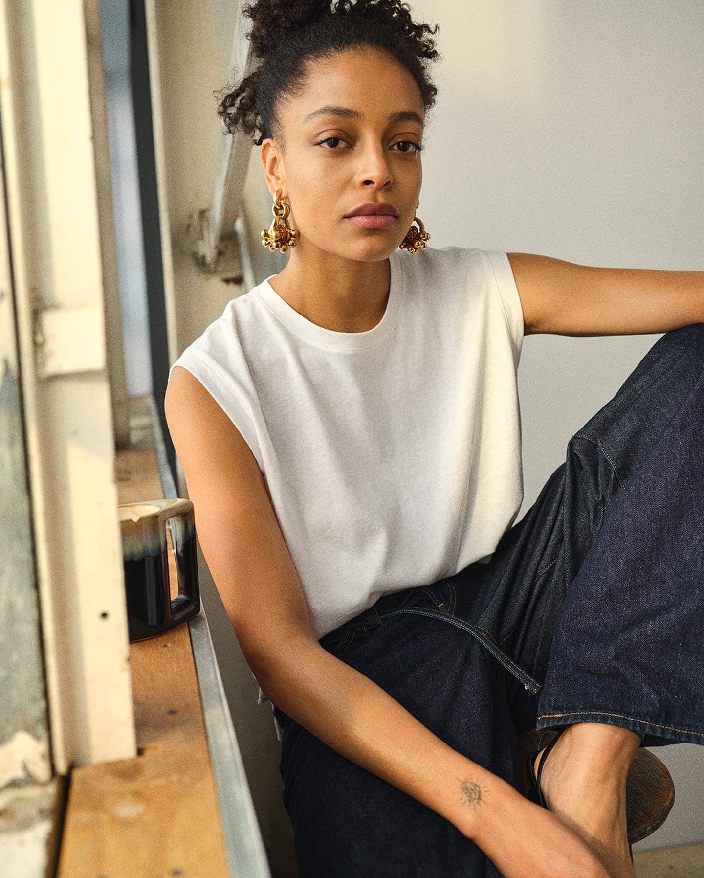 Portrait of a woman wearing a white sleeveless top and dark jeans seated indoors near a window.