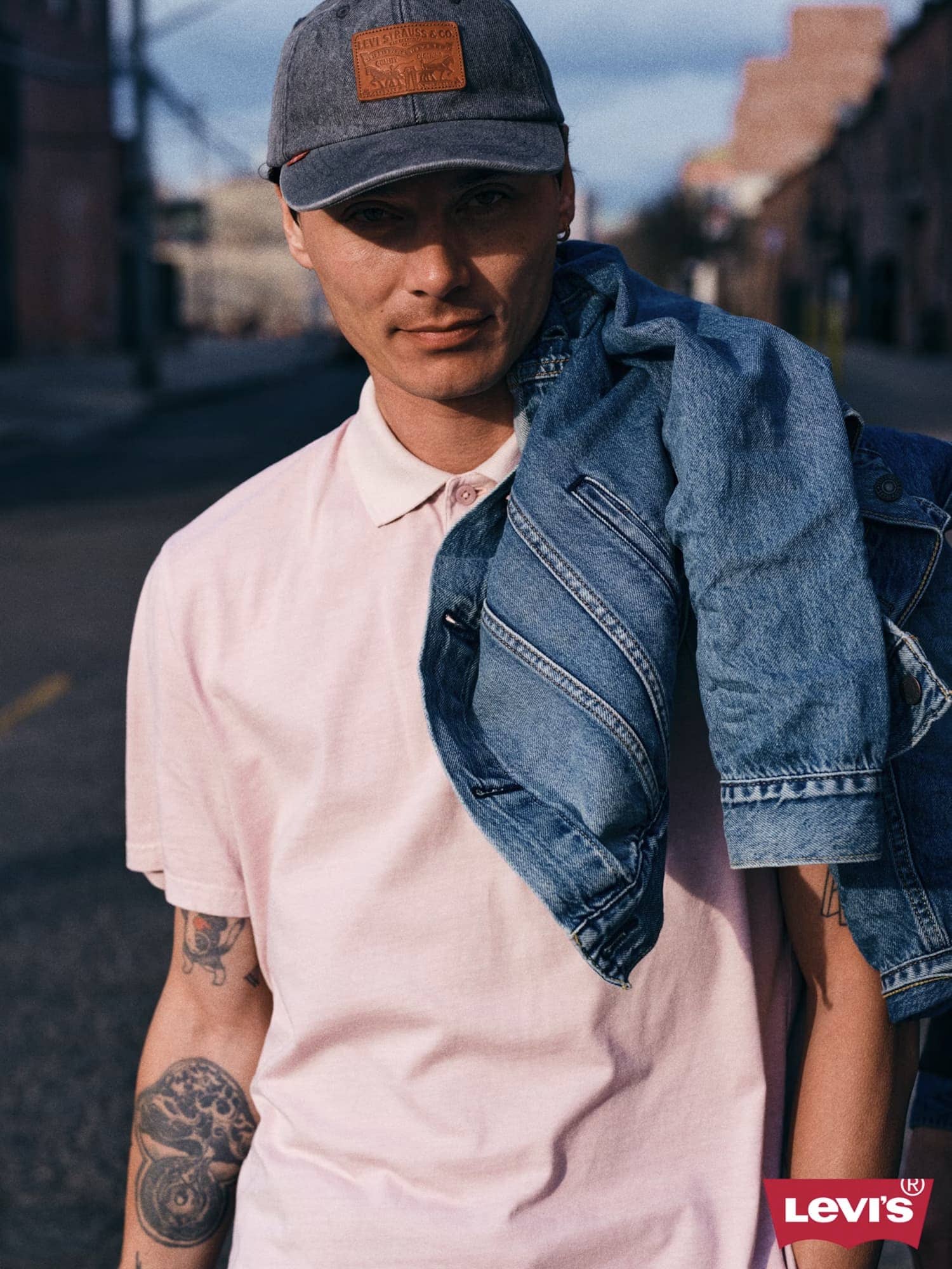Man wearing a pink polo shirt and grey baseball cap, holding a blue denim jacket over his shoulder on an outdoor street.