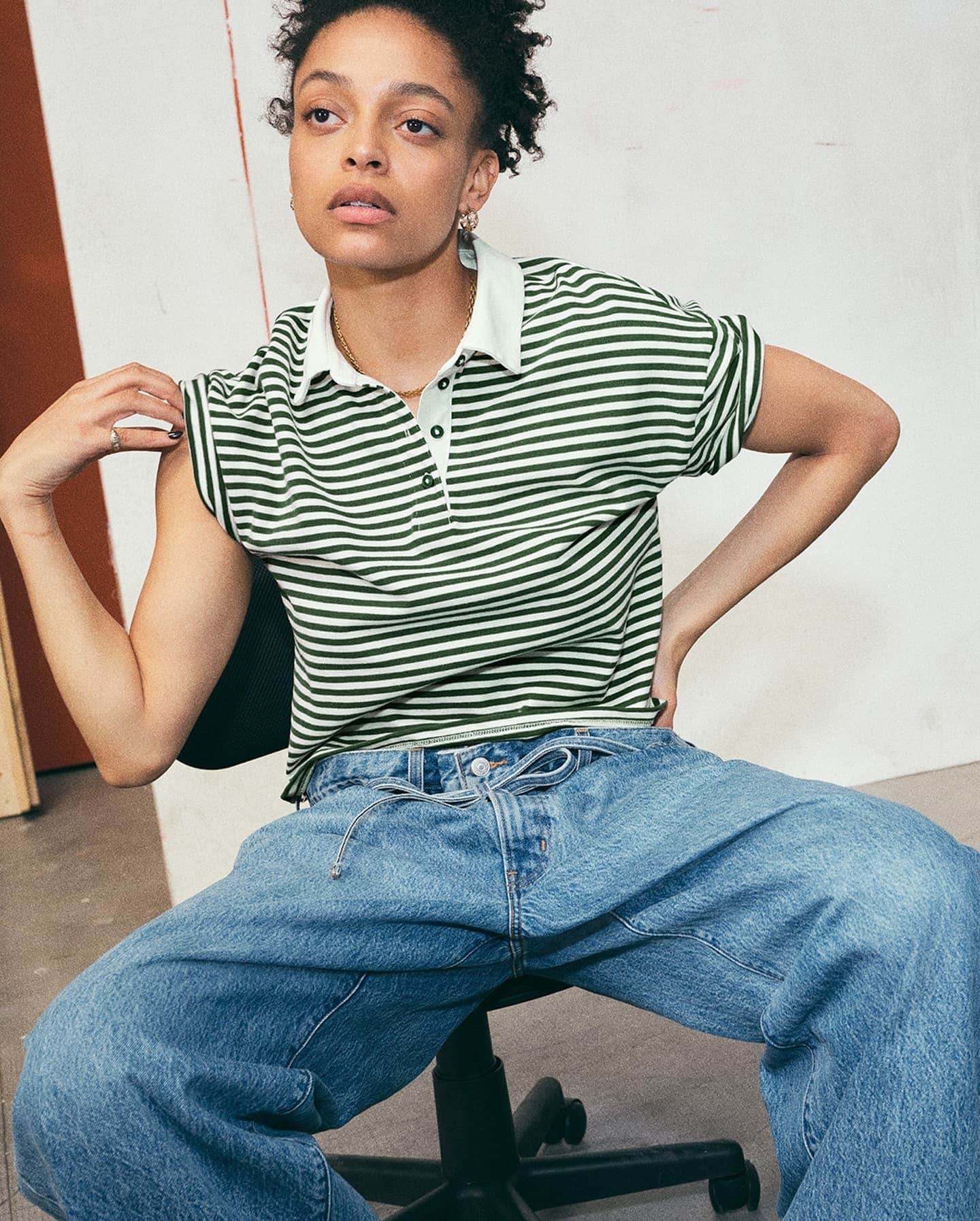 Woman in green and white striped polo shirt and loose denim jeans seated against a plain wall