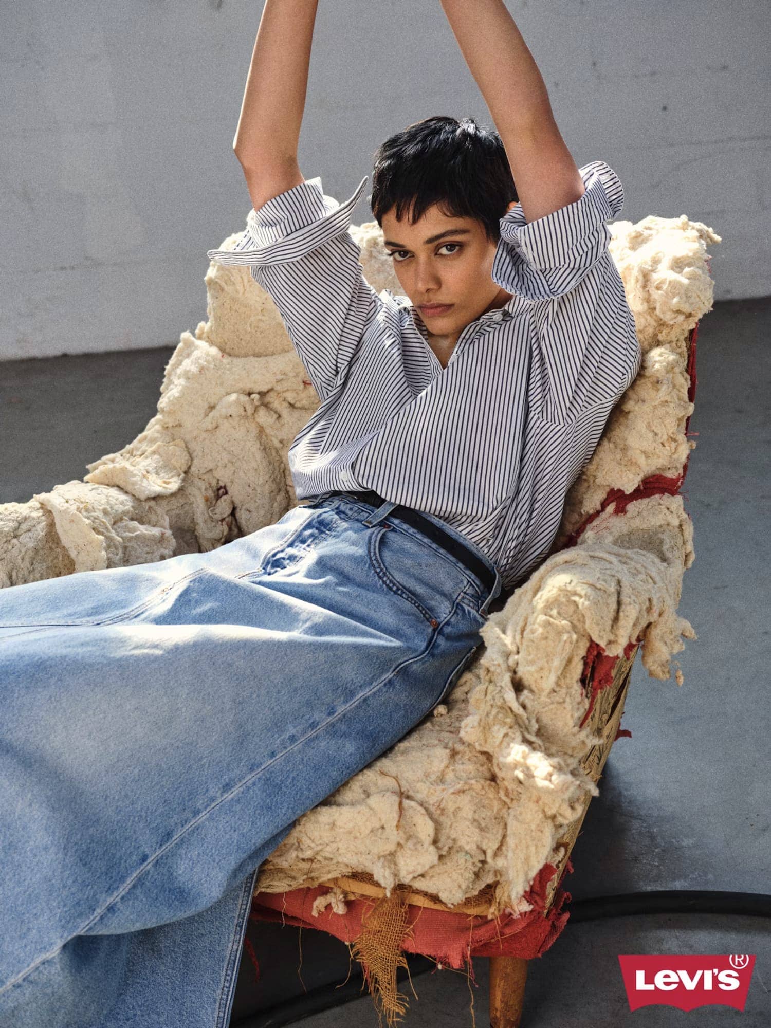 Model seated in an armchair wearing a striped shirt and denim skirt with arms raised for Levi's campaign