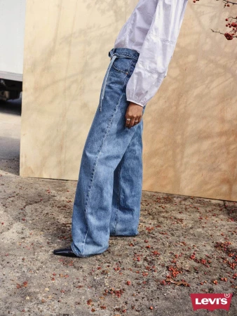 Person wearing wide-leg blue jeans and a white blouse standing outdoors near a wooden panel and red berries