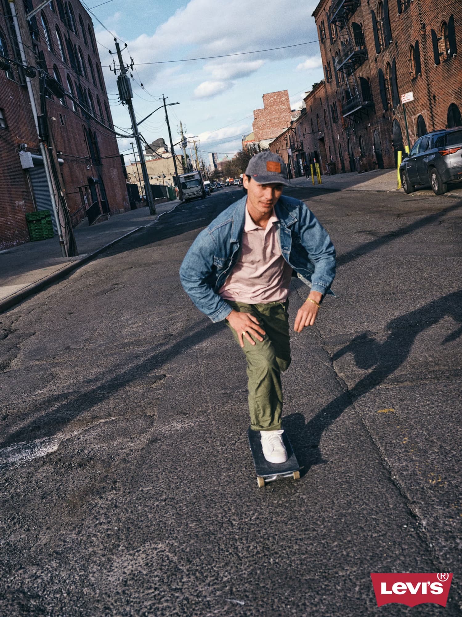 Man skateboarding down an urban street wearing a denim jacket, polo shirt, and baseball cap for Levi's campaign.