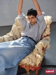 Model seated in an armchair wearing a striped shirt and denim skirt with arms raised for Levi's campaign