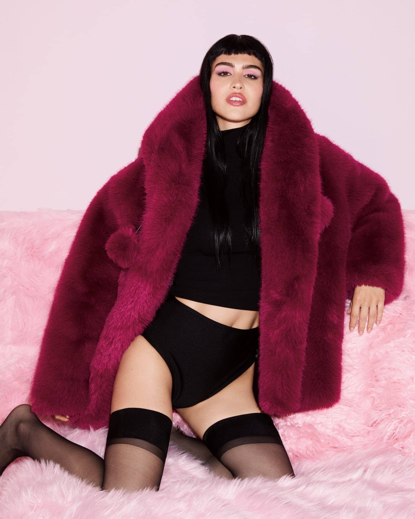 Model wearing a large burgundy furry coat over black apparel posed on a pink textured surface