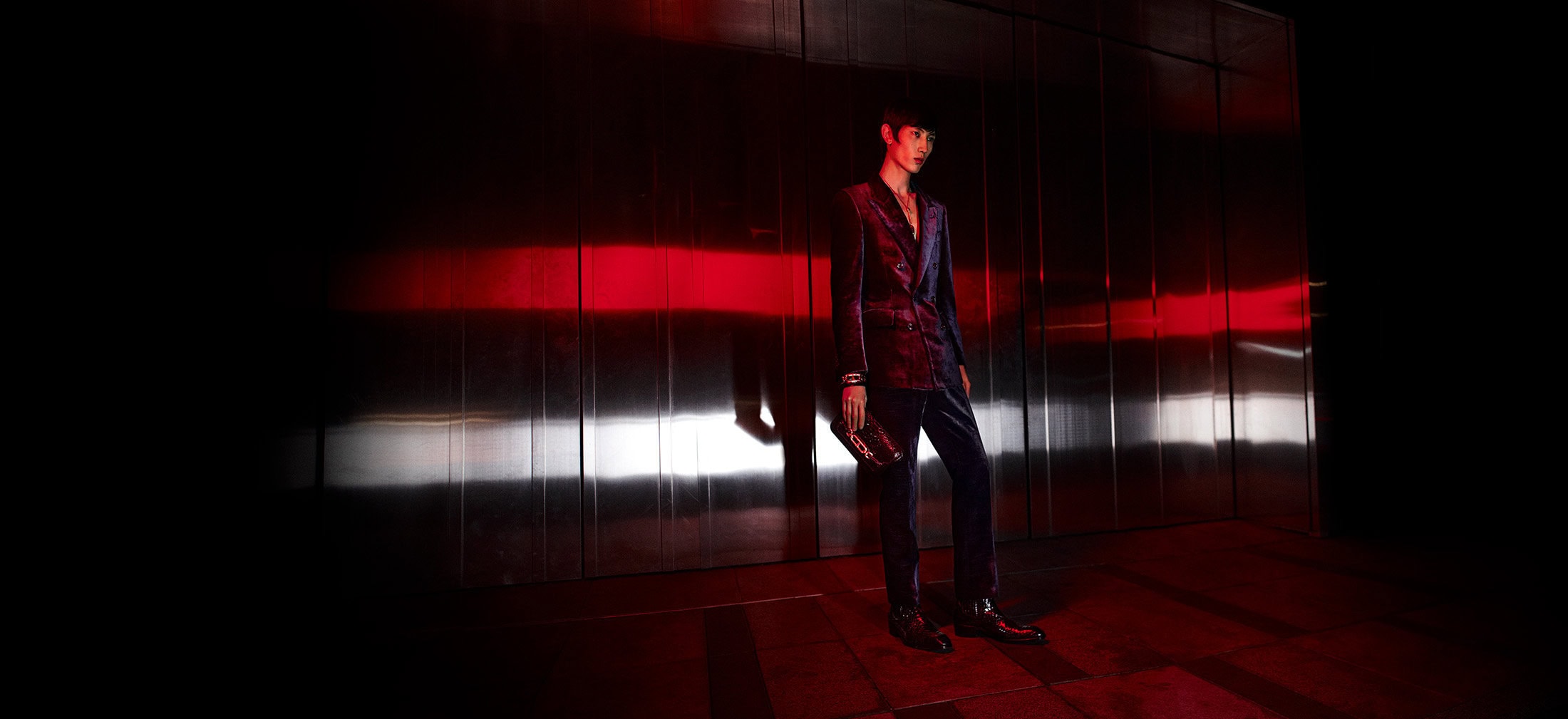 Man wearing a dark velvet suit holding a clutch against a gleaming metallic background with dramatic red light.