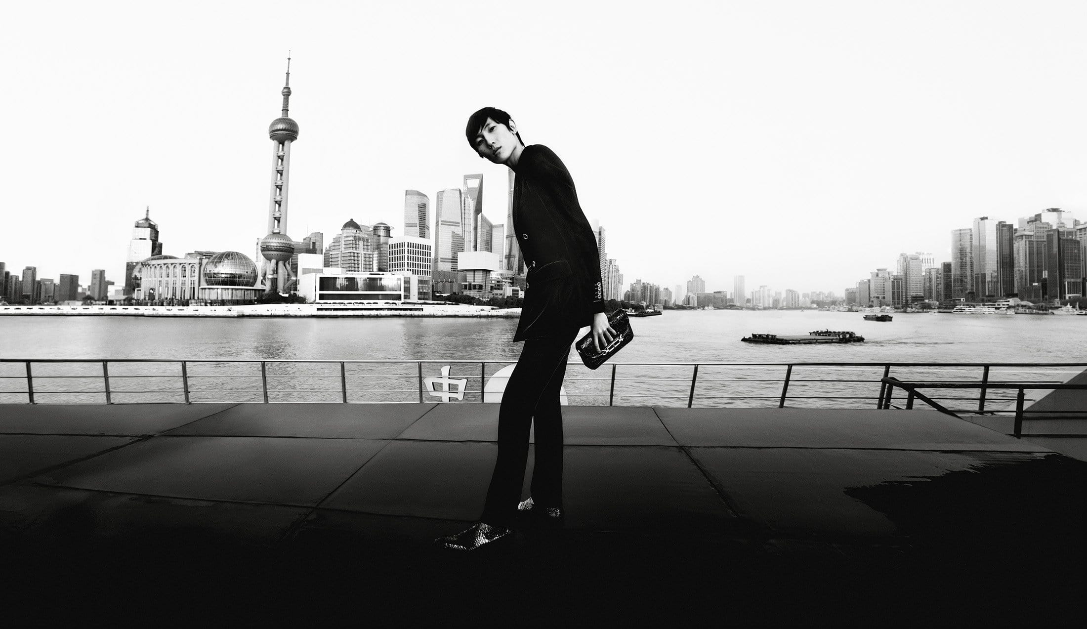 Model in dark tailored suit holding a handbag outdoors with city skyline across water in background