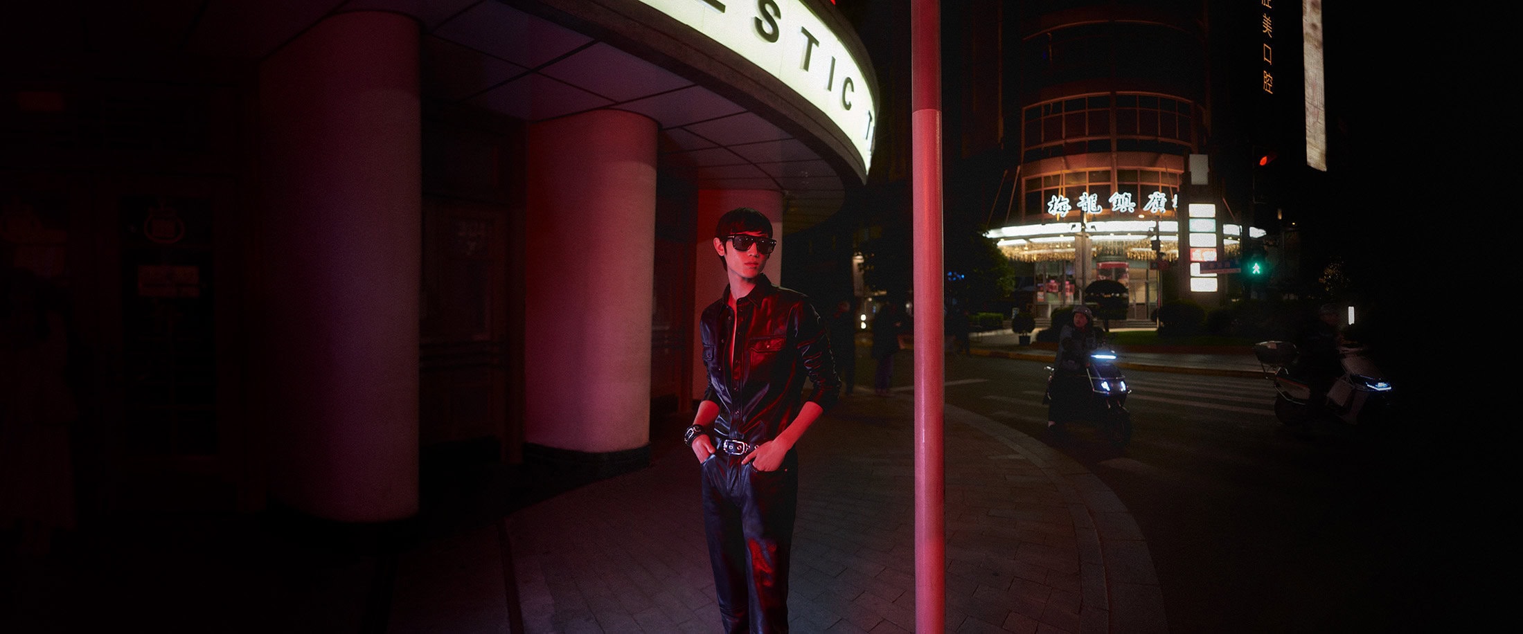 A man in black leather clothing and sunglasses standing on a nighttime city sidewalk under neon illumination for Tom Ford.