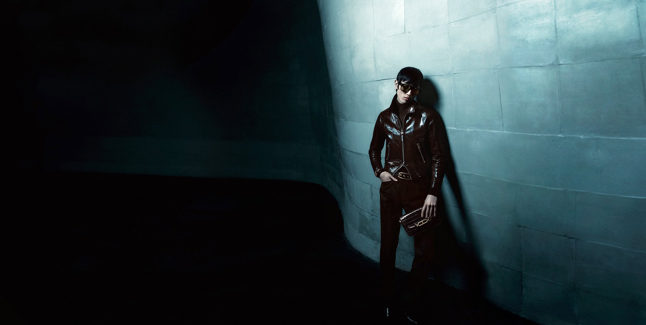 Model in dark sunglasses and brown leather jacket leaning against a curved tiled wall under low-key lighting for Tom Ford Winter 2024