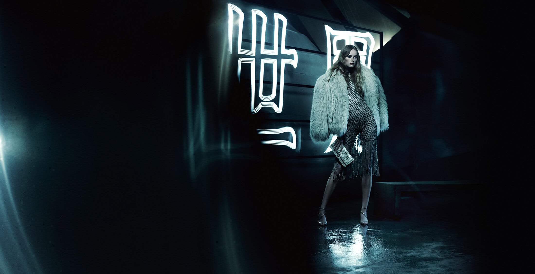 Model wearing a mesh dress and fur jacket standing before large neon Chinese characters in a dimly lit dark space