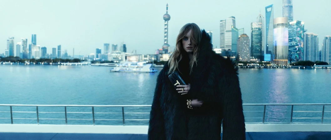 Model wearing a dark faux fur coat holding a black clutch in front of a Shanghai city skyline and river backdrop.
