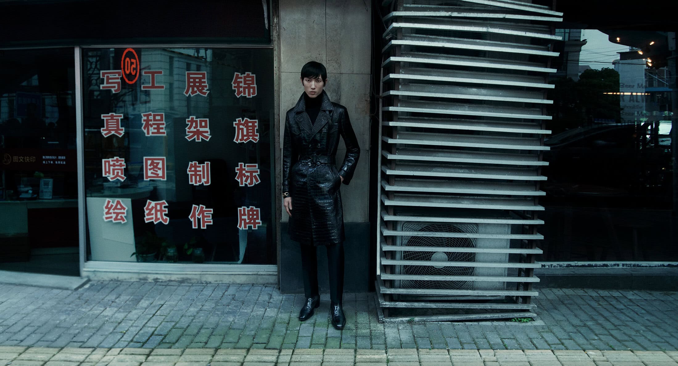 Man wearing a long black coat standing on a city sidewalk next to a storefront window with Chinese characters.
