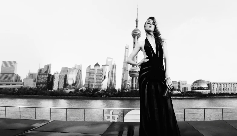 Model in a black evening gown standing along a waterfront with a cityscape and tower in the background.