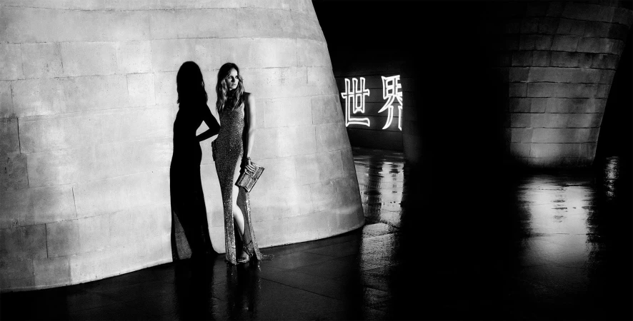 Monochrome fashion campaign image featuring a woman in a shimmering gown holding a clutch against a stone pillar.