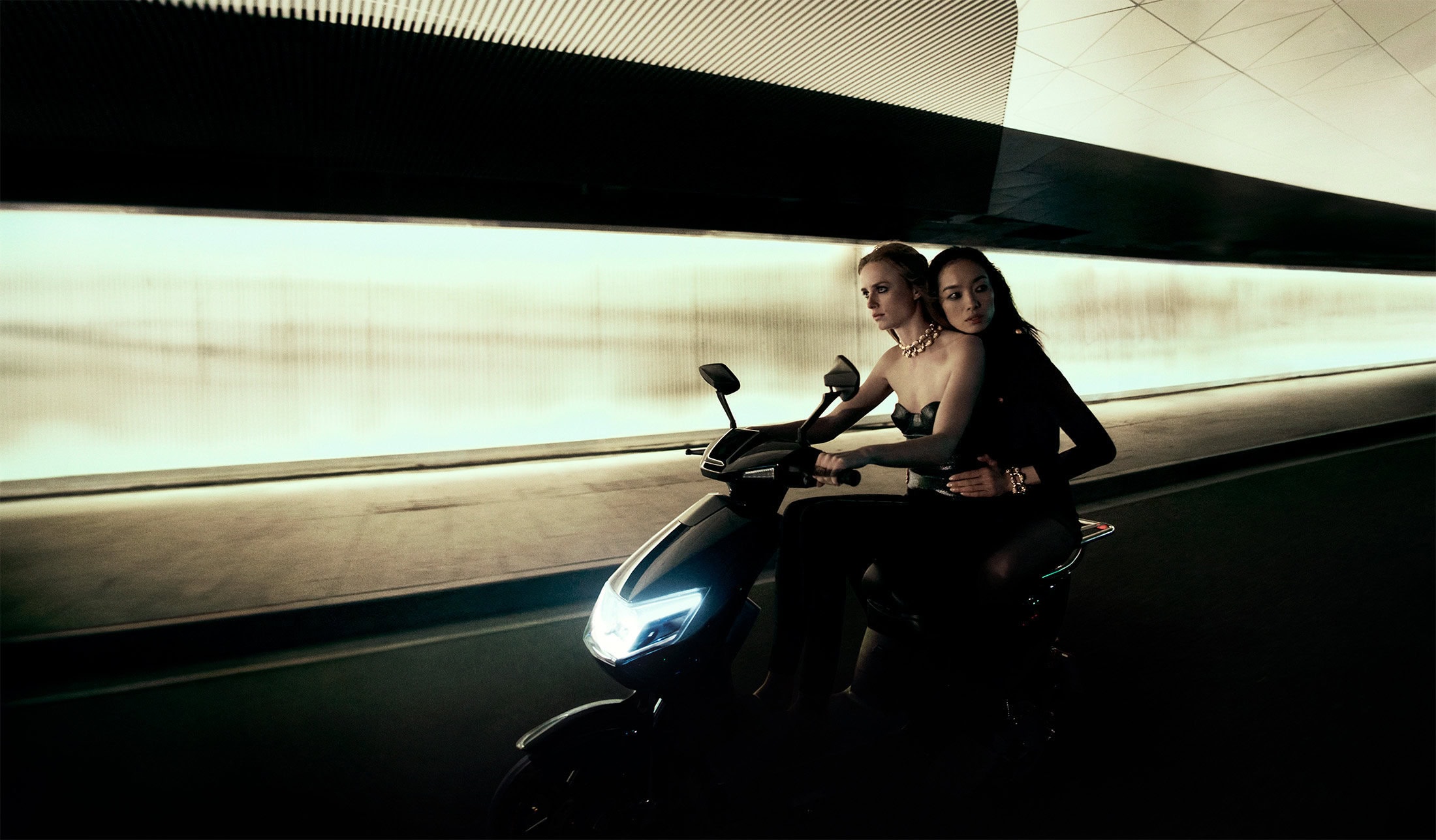 Two women riding a modern black motor scooter through a brightly illuminated highway tunnel at night.