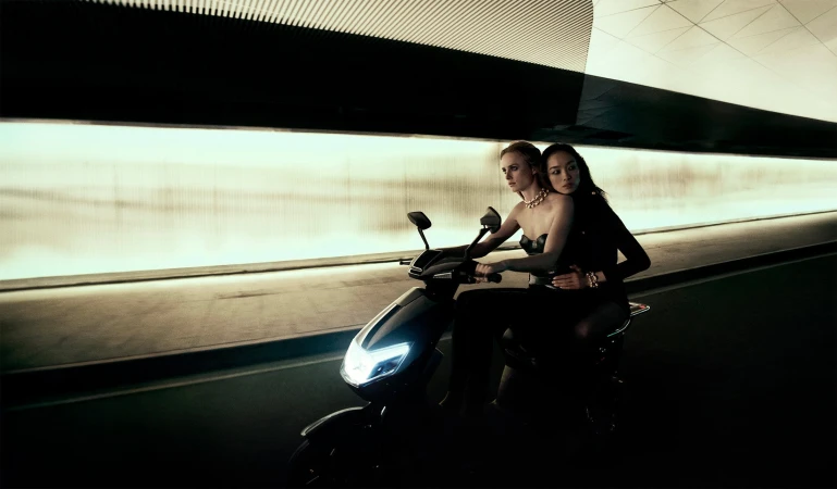 Two women riding a modern black motor scooter through a brightly illuminated highway tunnel at night.