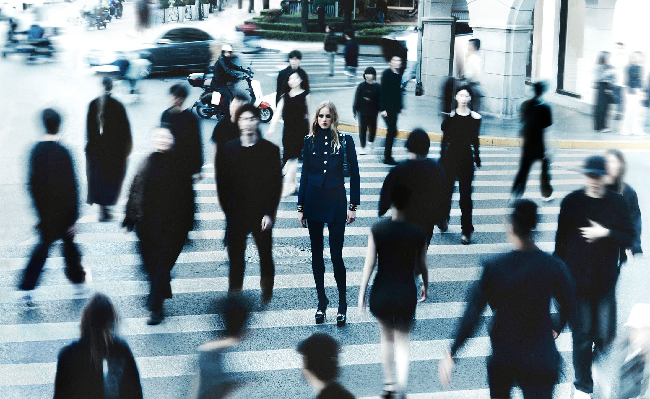 Model standing still in a dark blue military-style jacket amid blurred pedestrians crossing a city street