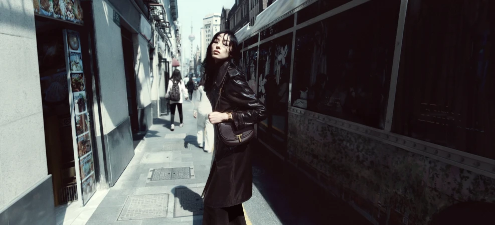Woman in long dark leather coat holding a shoulder bag on a sunny city street