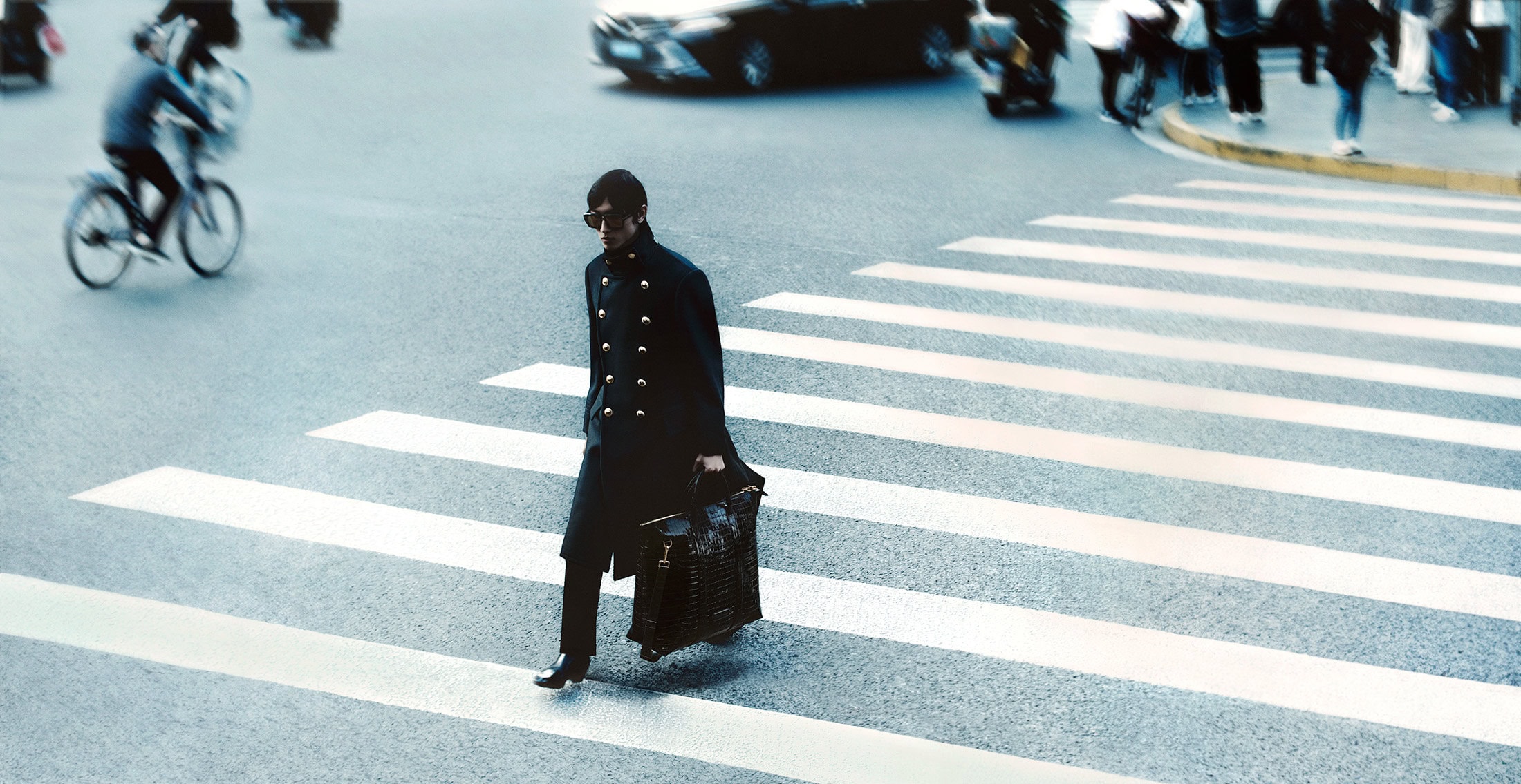 Man wearing dark coat and sunglasses walking across a zebra crossing carrying a large bag