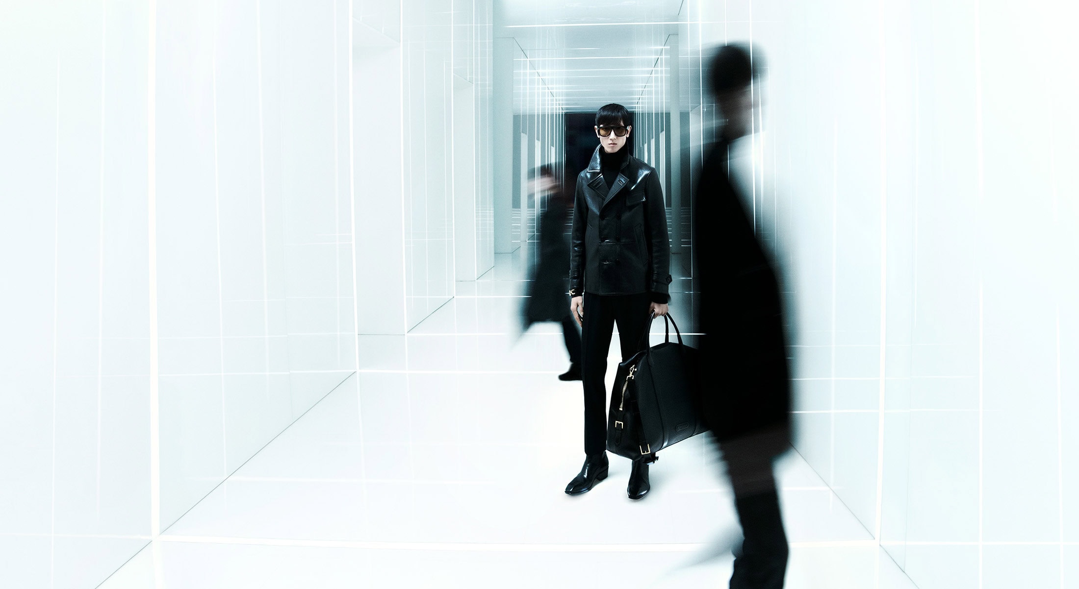Man in dark leather jacket and trousers standing in a bright white corridor with blurred figures in motion