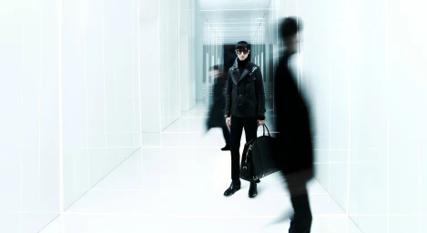 Man in dark leather jacket and trousers standing in a bright white corridor with blurred figures in motion
