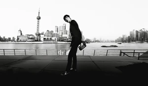 Model in dark tailored suit holding a handbag outdoors with city skyline across water in background