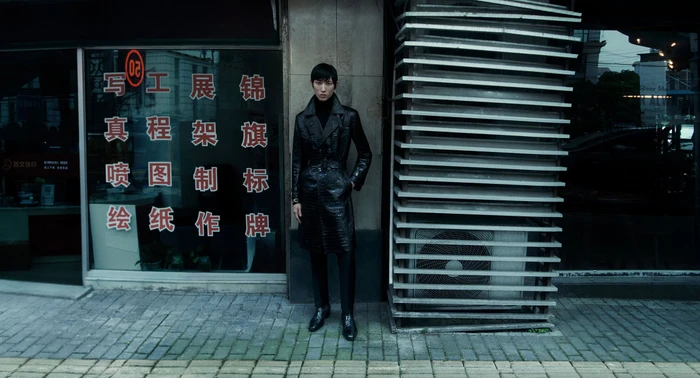 Man wearing a long black coat standing on a city sidewalk next to a storefront window with Chinese characters.