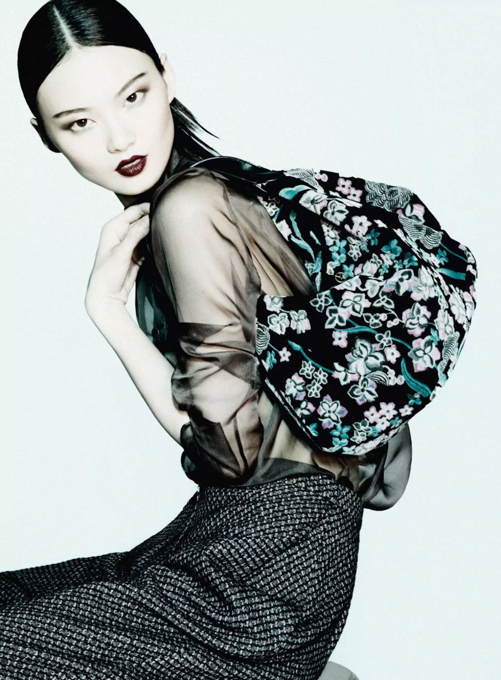 Model with dark hair and dark lipstick wearing a sheer top and patterned floral backpack against a bright white background